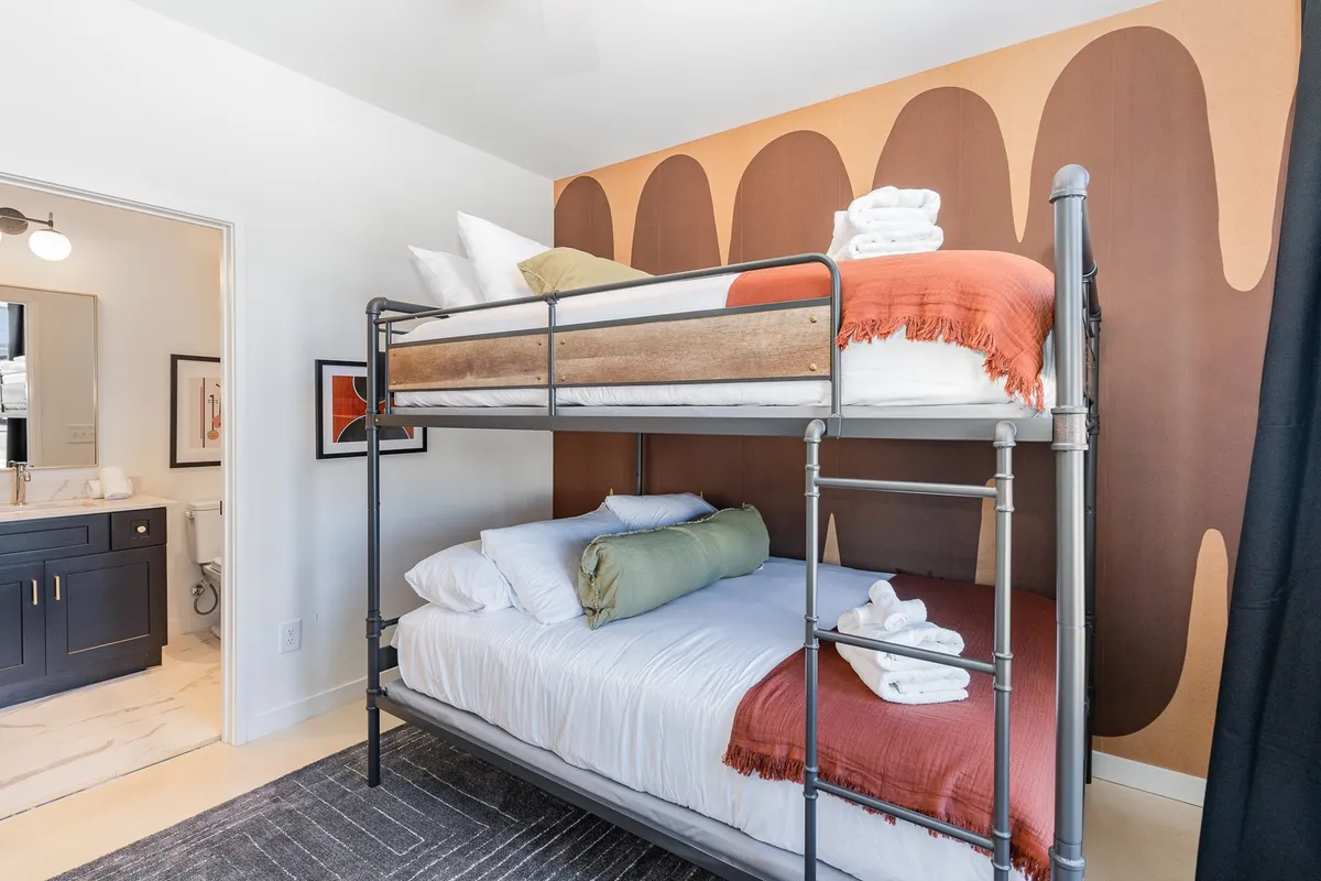 A cheerful bunk room is featured here, with a fun bunk bed setup made up of two queen beds, colorful wall art, and the entrance to an adjacent bathroom visible. The ambiance is playful and inviting, ideal for group stays or families with kids.