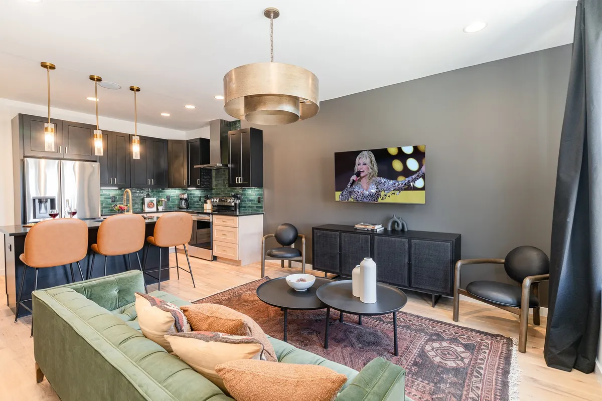 A wide view of the open-concept living area, featuring a luxurious green velvet sofa, sleek black kitchen cabinetry, a large wall-mounted TV, and stylish pendant lighting. The scene includes both kitchen and living spaces, emphasizing the room's unity and sociable layout.