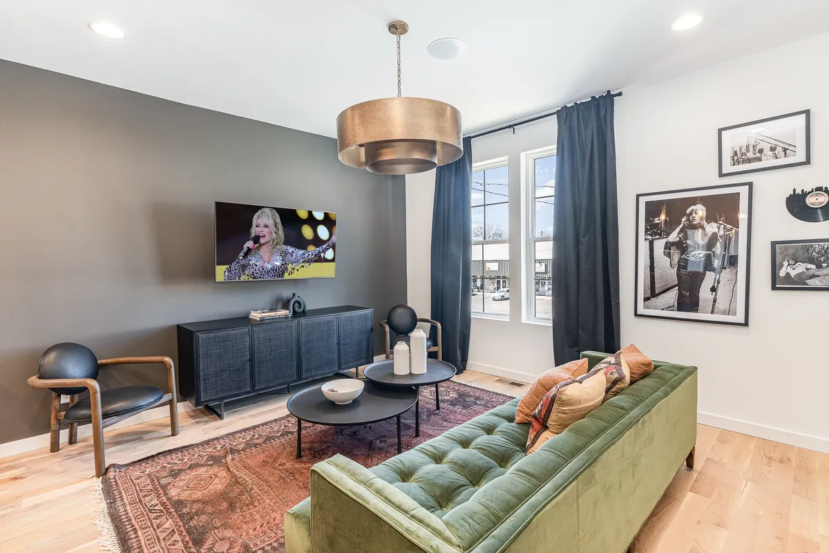A cozy yet stylish living space, anchored by a green tufted sofa, black media console, and large television displaying a live performance. The room is decorated with framed photographs and a round coffee table, suggesting evenings spent relaxing or watching movies.
