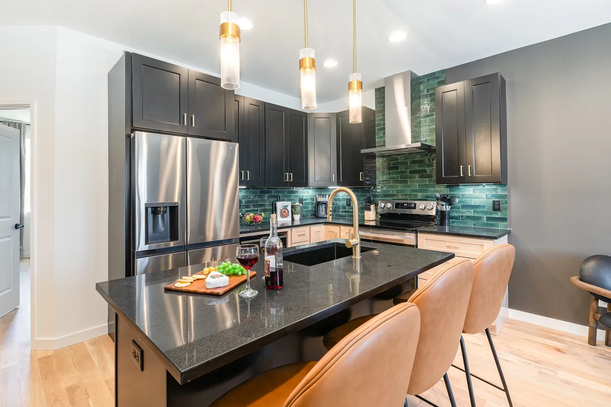 A vibrant kitchen island with bar stools, shining stainless steel appliances, and an eye-catching green tile backsplash. The space is set up for friendly gatherings and culinary adventures, with ample counter space and room for socializing while cooking.