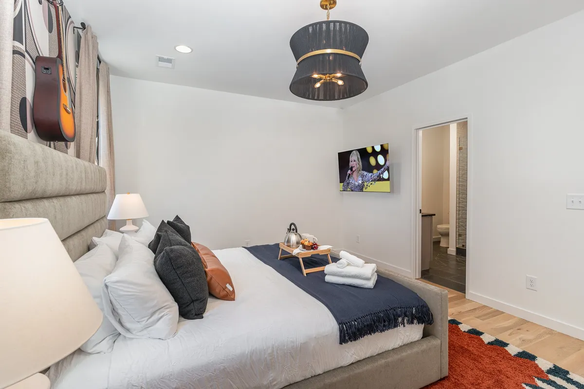 This image shows the primary bedroom featuring a king-sized bed adorned with decorative pillows. A TV is mounted in front of the bed, and a breakfast tray suggests leisurely mornings. Unique guitar decor on the wall adds a Nashville touch and a sense of local character.